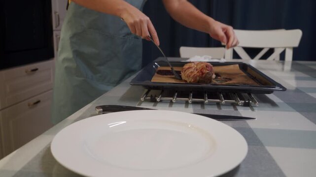 A woman wearing an apron uses a spatula to lift hot bacon-wrapped meat from a baking tray. She prepares to serve the freshly cooked dish onto a white plate on the kitchen table.