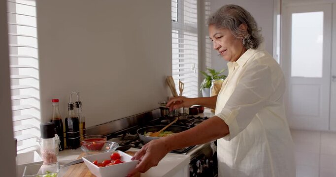 Elderly senior woman cooking vegetables in kitchen, smiling while adding spices to pan