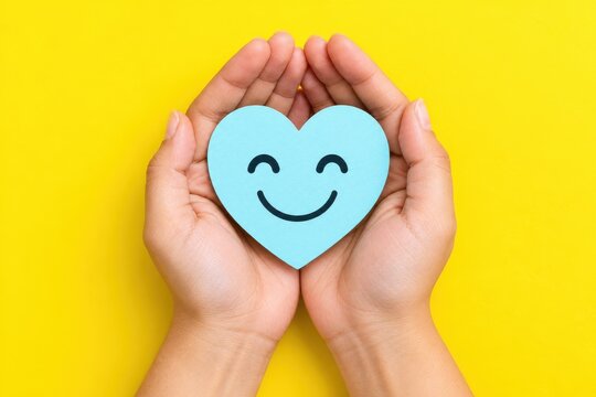 Two hands cradle a light blue heart with a cheerful smile during World Smile Day against a vibrant yellow background