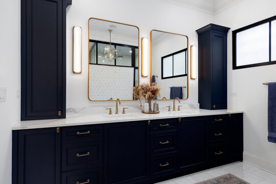 A luxury bathroom with dark blue cabinet, gold faucets on a white marble countertop, gold sconces and mirrors, and tile flooring. 