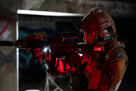 A tactical operator aims a rifle indoors under low light with red glow casting shadows, conveying a sense of intense focus, vigilance, and strategic readiness during a mission.