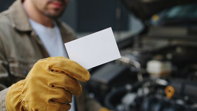 Mechanic wearing yellow leather work gloves holding a blank white business card in front of an open car engine hood in a garage workshop