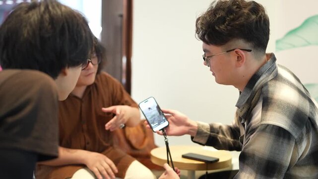 A man in a plaid shirt holds a smartphone toward a seated companion so the screen can be viewed.