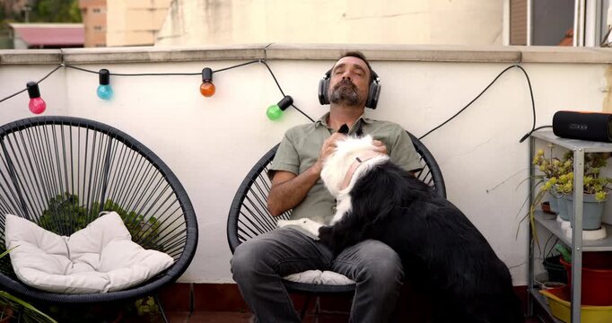 Relaxed man with headphones and dog on balcony: A man with headphones on, enjoying music on his balcony, is nuzzled by his loyal dog. Outdoor relaxation setting with string lights and plants.