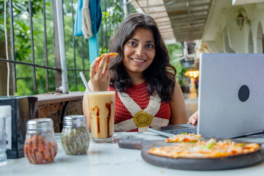 Young indian gen z woman working on laptop while enjoying a snack and a coffee milkshake at a restaurant or cafe, Concept of modern digital nomad lifestyle and remote work flexibility, freelancing