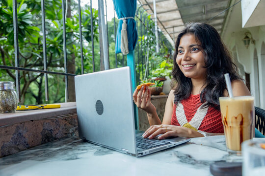 Young indian gen z woman working on laptop while enjoying a snack and a coffee milkshake at a restaurant or cafe, Concept of modern digital nomad lifestyle and remote work flexibility, freelancing