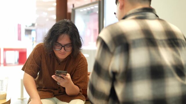 A man in a brown shirt smiles while looking at a smartphone as another man sits in the foreground.