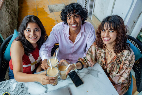 Young indian Gen Z friends enjoying a cheerful moment, clinking glasses of iced coffee at an outdoor cafe, celebrating friendship and social connection during a summer day