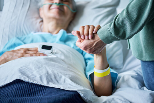 Close up of adult holding hand of senior Caucasian patient lying in hospital bed, offering emotional support. Concept of healthcare and palliative care marketing