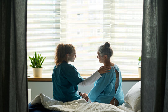 Young Caucasian female nurse comforting senior Caucasian female patient sitting on hospital bed. Visual metaphor for compassionate eldercare, nursing support and healthcare advertising