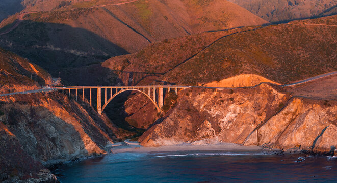 Aerial shot reveals Bixby Creek Bridge along the rugged Big Sur coast in California during sunset, where mountains meet the Pacific Ocean beach and highway.