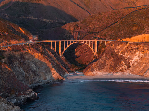 Aerial perspective of Bixby Creek Bridge along the Pacific Coast Highway in Big Sur California during sunset, showing coastal cliffs, the beach below, and ocean waters.