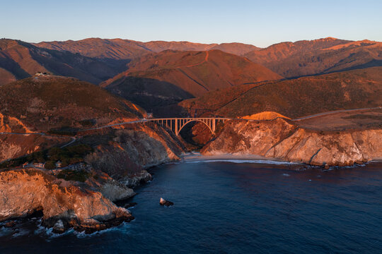 Aerial perspective displays Bixby Creek Bridge spanning a coastal canyon along Highway 1 in Big Sur, California, surrounded by ocean waters, cliffs, and sunlit mountains during sunset.