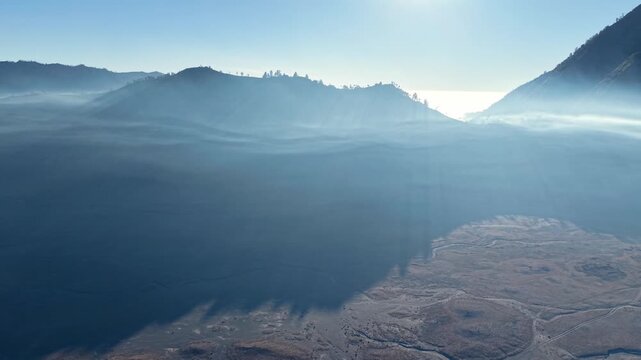 Mist drifts over calm lake, hills rise softly behind. Sunlight pierces mist, casting slow near Bromo