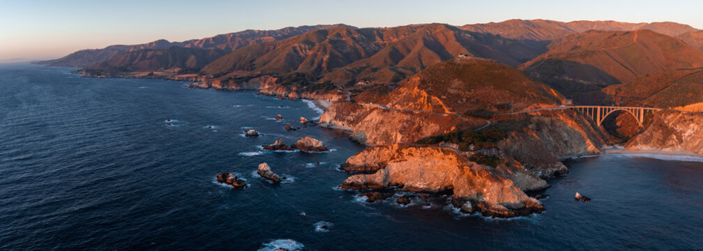 Aerial panoramic view captures Bixby Creek Bridge spanning a canyon along the Pacific coastline of Big Sur California at sunset, showing warm light on cliffs and hills.