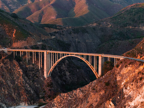 Aerial shot depicts Bixby Creek Bridge spanning a steep coastal canyon in Big Sur, California during warm sunset illumination on the concrete arch and rugged mountains.