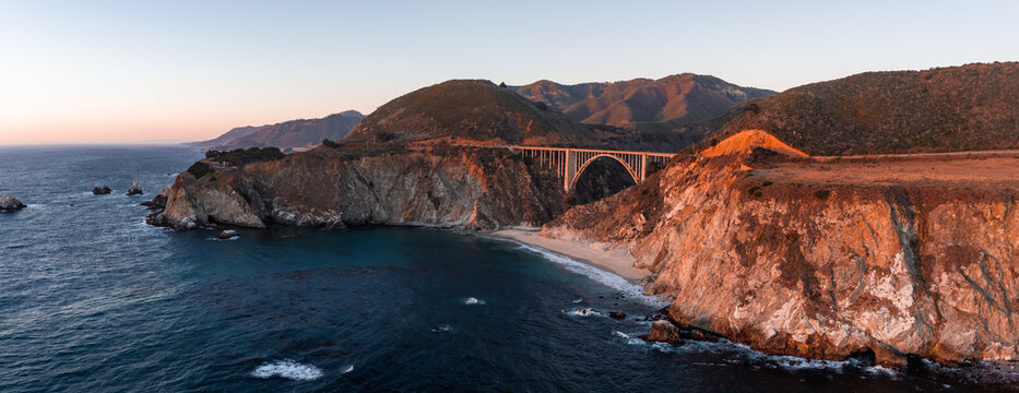 Aerial view showcases Bixby Creek Bridge spanning the rugged coastline of Big Sur in California during sunset, where cliffs drop down to the Pacific Ocean shoreline.