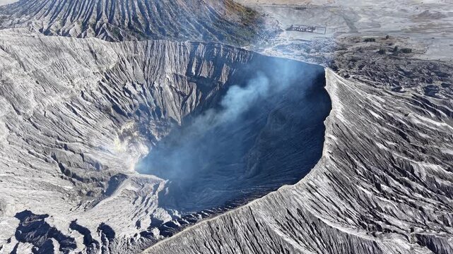 Aerial view of a smoking volcanic Bromo crater. Rugged cliffs surround the dark, steaming bowl