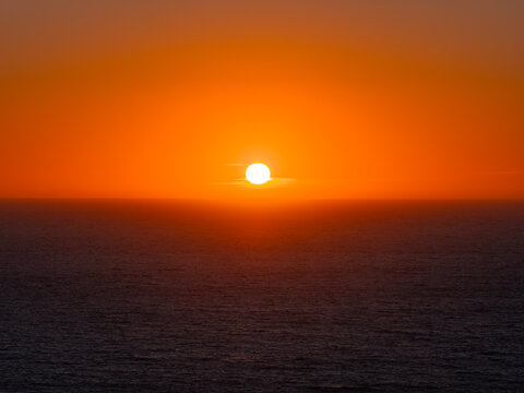 Bright sun sets above the horizon of the Pacific Ocean along the California coast at dusk. The sky shines with intense orange light over open dark sea waters.