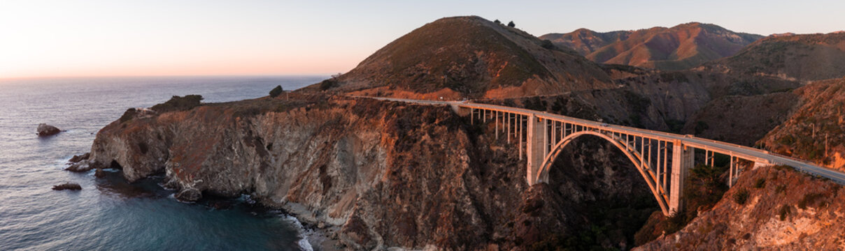 Aerial panorama of Bixby Creek Bridge illuminated by the warm evening light along the rugged coastal cliffs and ocean waters of Big Sur, California during dusk.