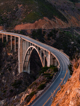 Bixby Creek Bridge curves along Highway 1 in Big Sur, California, spanning a deep coastal canyon flanked by rocky cliffs and natural vegetation at dusk.