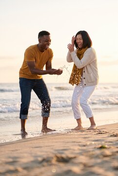 Playful couple splashing seawater barefoot together on sunlit sandy beach shore
