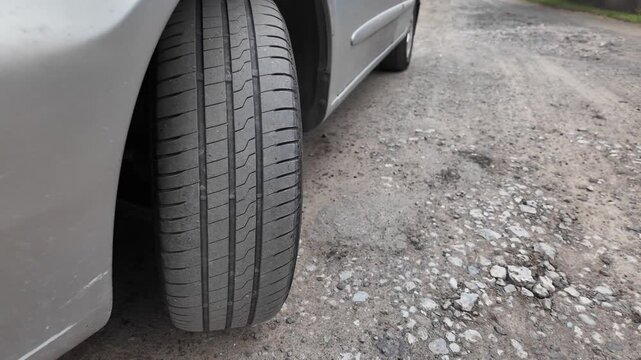 Close-up shot of a car tire resting on a dirt and gravel road. The detailed view highlights the rubber tread pattern and the edge of a grey vehicle bumper. Great for auto maintenance concepts.