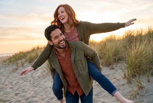 Playful couple piggyback riding with arms outstretched on golden beach dunes