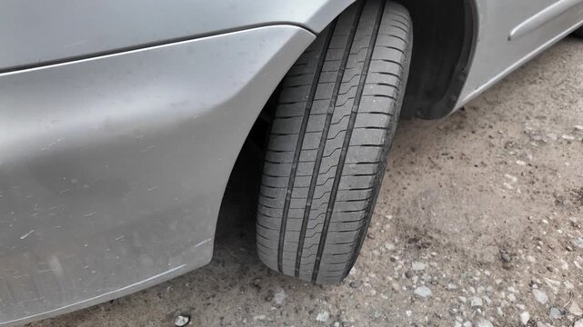 Close-up shot of a car tire resting on a dirt and gravel road. The detailed view highlights the rubber tread pattern and the edge of a grey vehicle bumper. Great for auto maintenance concepts.