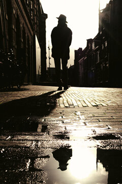 Silhouetted man in hat walking on cobblestone street with puddle reflection at sunset