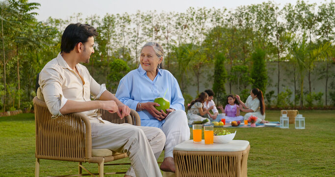 Indian old aged mom sitting on chair with young adult son talk share gossip hold fresh coconut drink juice outdoor Happy male man older female speak funny spend time enjoy picnic trip day garden place