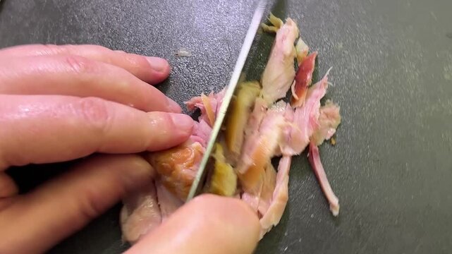 Hands use a broad kitchen knife to separate cooked chicken meat from the bone beside sliced pieces on a cutting board.