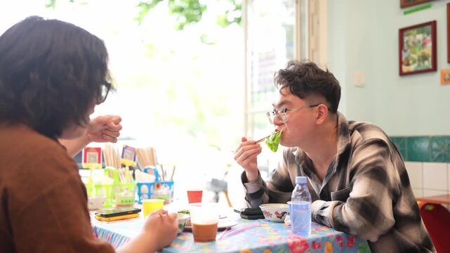 A man in a plaid shirt leans over the table and eats food with chopsticks.