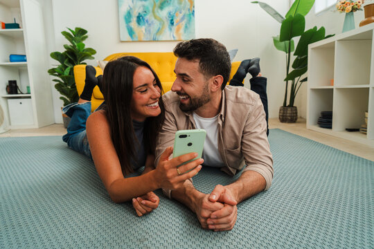 Two people enjoy a relaxed moment on a soft rug, exploring content on a shared phone. Their warm smiles and close posture convey connection, curiosity, and lighthearted teamwork during a casual home