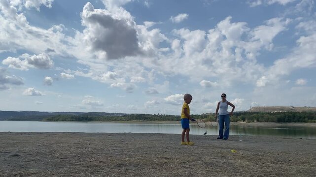 A happy young boy walks with a badminton racket as his mother stands behind him. They enjoy playing on a dry shore by a quiet lake under a cloudy sky. Their fun moment feels warm and peaceful.