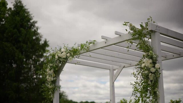 Elegant white wooden wedding pergola decorated with fresh flowers and lush green foliage outdoors