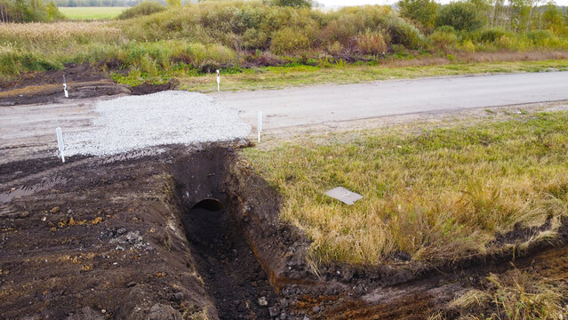 An elevated outdoor photograph capturing a drainage culvert pipe installed inside a newly excavated dirt trench running perpendicular to a dirt road.