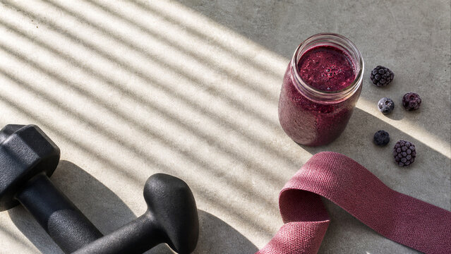 Berry smoothie in glass jar with dumbbell and resistance band on gray background. Healthy food and fitness equipment flat lay with sunlight shadows.