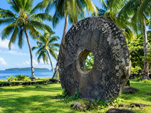 Giant carved Yap stone money disc stands on grass under palm trees in bright daylight