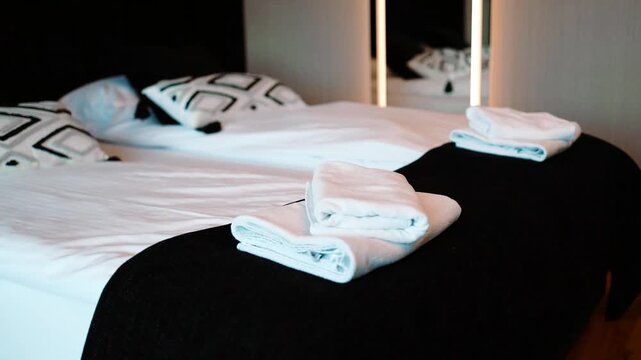  Close-up of a neatly made double bed in a modern hotel room, featuring white linens, decorative pillows, dark blankets, and rolled clean towels prepared for guests.