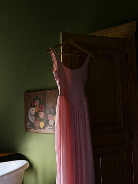 Pink vintage style dress on wooden hanger hanging on antique wardrobe door in dark olive green room, moody natural light