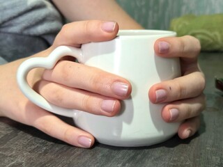 Hands holding white mug on table. Close-up of hands with manicure holding a white ceramic mug on a...