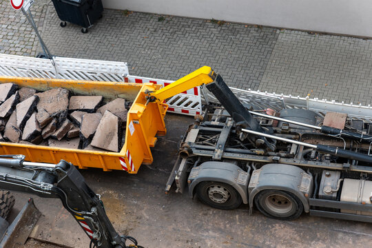Hooklift truck loads broken asphalt and concrete into roll off container during urban roadwork. Excavator, rubble and heavy machinery show demolition, recycling and infrastructure repair process