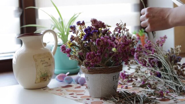Woman arranging colorful dried statice bouquet in rustic pot near window, cozy slow living floral DIY craft