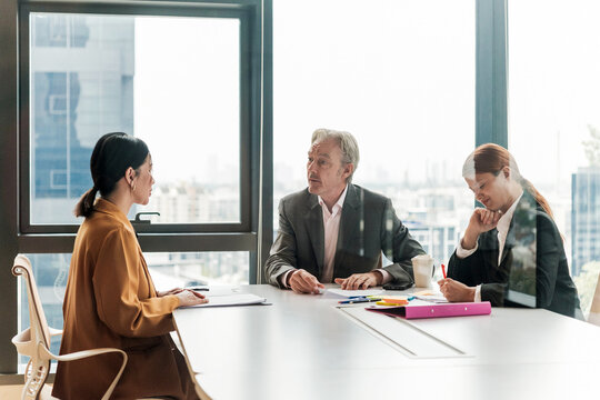 Three professionals in a modern office hold a formal meeting, likely a job interview, as one candidate speaks and two interviewers take notes by large windows with a city view.