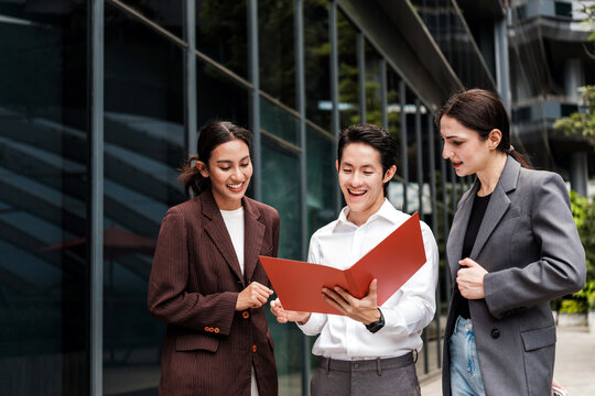 Three professionals outside a modern office review a red folder, smiling and collaborating on a project beside glass building facades.