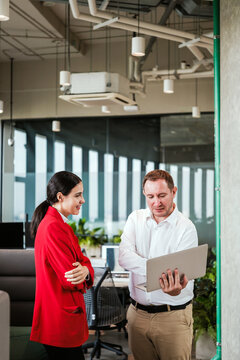 Two coworkers discuss a project in a modern open office, reviewing a laptop amid glass walls and ceiling lights.