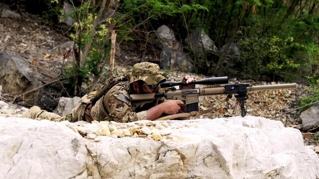 Camouflaged sniper lying prone behind rocks in a forested area, aiming a suppressed, scoped rifle during a tactical operation or training