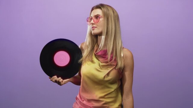 Woman holding a black vinyl record with pink label and wearing pink heart sunglasses in studio, posing with head turned and relaxed casual gaze; nostalgia retro music.