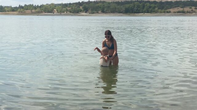 A caring mother gently guides her young son wearing a swim vest in the water. They enjoy a refreshing dip together in a clear, calm lake. Their sweet bonding time feels warm and safe.